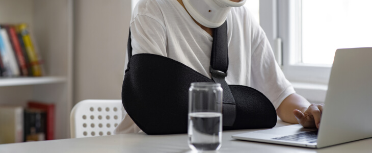 Woman wearing neck brace and arm sling working on a laptop