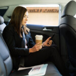 Busy businesswoman drinking coffee to go in the back of a taxi cab while sending a text on her smartphone.